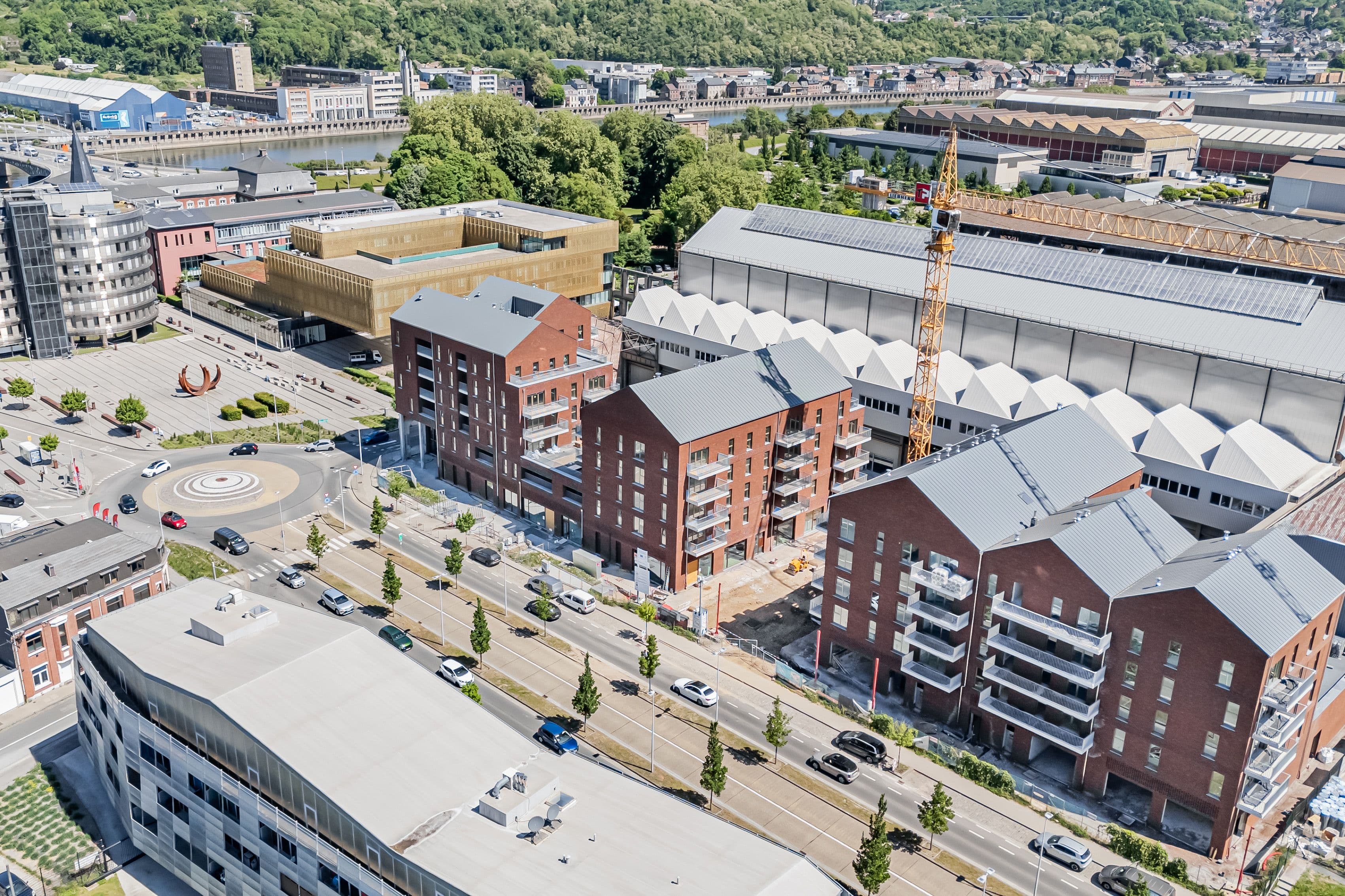 Vue aérienne d’un projet résidentiel en cours de construction, composé de plusieurs immeubles en briques avec balcons. Le site s’inscrit dans un environnement urbain mixte, à proximité d’un rond-point, d’axes routiers et d’une rivière.